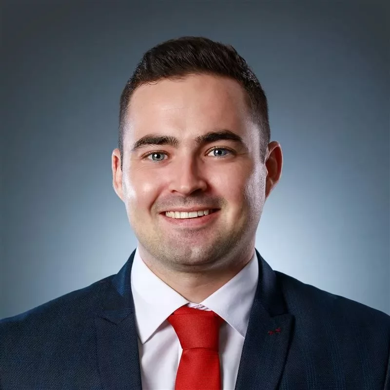 Person in a suit with a red tie smiling against a blue background.