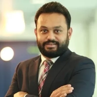 Smiling person with a beard in a suit and checkered tie, standing with arms crossed in an office setting.