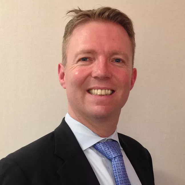 Person smiling in a suit and patterned tie against a plain background.