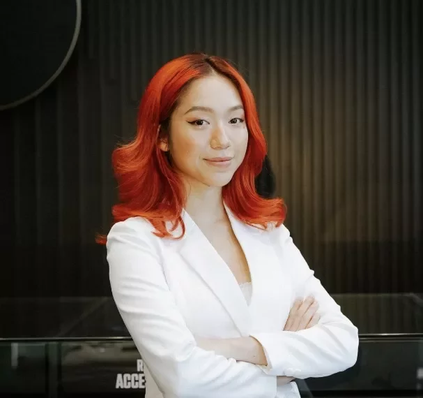 Person with red hair in a white outfit stands confidently with arms crossed.