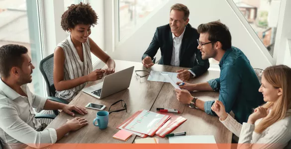 A group of people having a meeting at a wooden table with papers and a laptop.