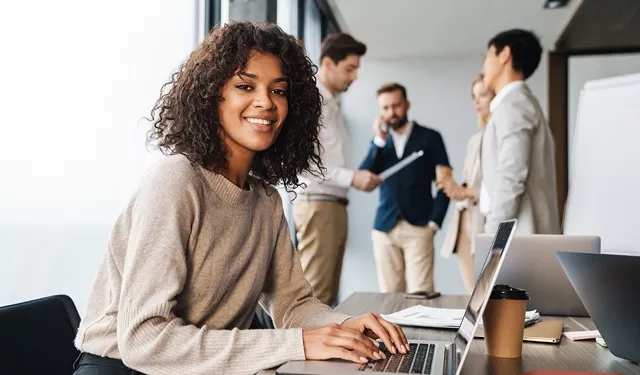 Person smiling at a desk with laptop, group talking in background.