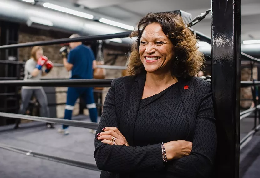 Smiling person in a suit stands by a boxing ring with boxers in the background.