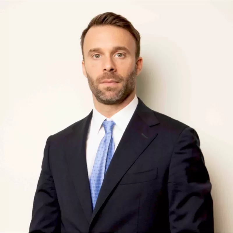Person in a suit with a blue tie standing against a neutral background.