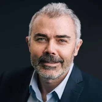 Portrait of a smiling person with short gray hair and a beard, wearing a suit and light shirt.