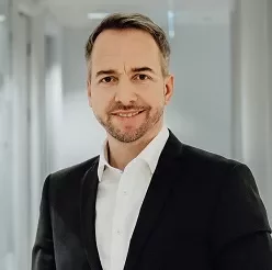 Smiling person in a suit standing in a bright hallway.