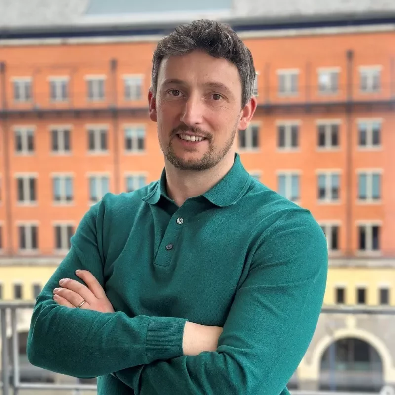 Person in green shirt smiling in front of a brick building.