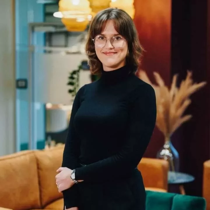 Person wearing glasses and a black outfit stands in a cozy room with warm lighting.