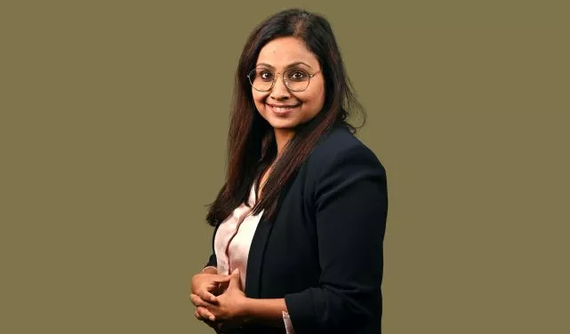 Individual in a blazer and glasses smiles with hands folded.