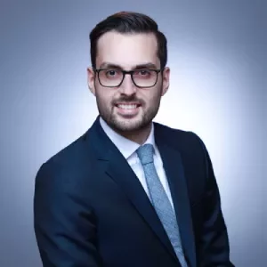 Person in a suit and tie with glasses, smiling against a blue backdrop.