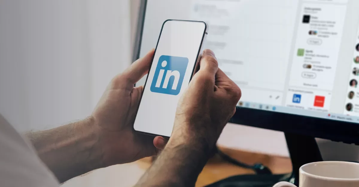 Hands holding a smartphone displaying the LinkedIn logo, with a computer screen in the background.