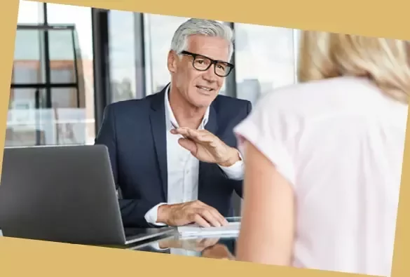 Person speaking with another person across a desk in an office, with a laptop in the foreground.