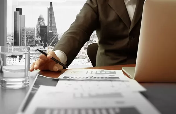 Person in a suit working at a desk with a laptop and papers, cityscape visible through the window.