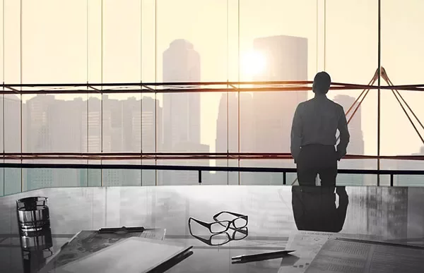 Person looking at city skyline through large office window, desk in foreground.