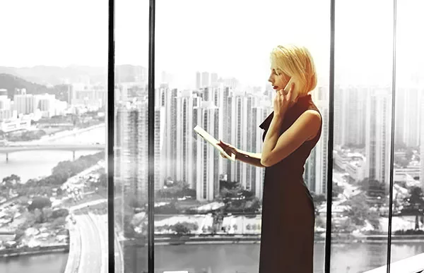 Person standing by large window, holding a tablet with a cityscape in the background.