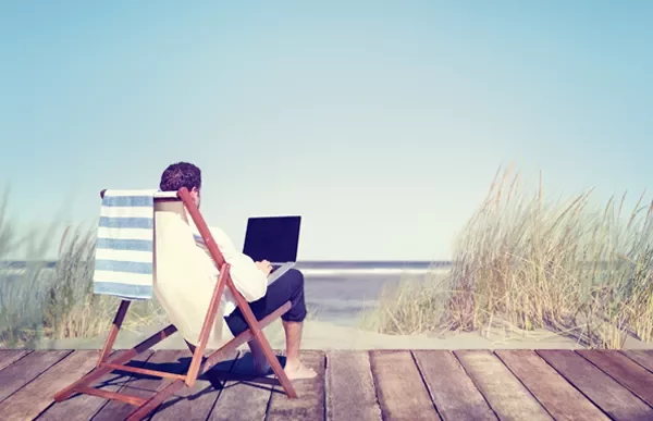 Person relaxing on a deck chair by the beach with a laptop.