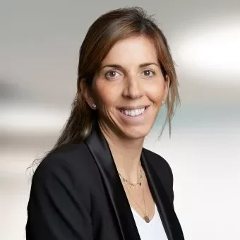 A person smiling, wearing a black blazer and white shirt, blurred background.
