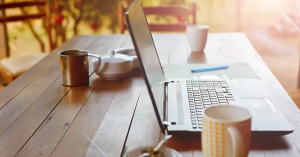 Open laptop on a wooden table with mugs and a teapot in a sunlit setting.