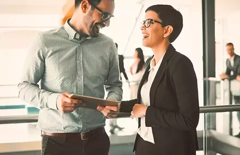 Two colleagues laughing while discussing work with a tablet in an office setting.